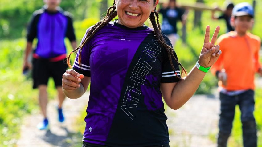 A cheerful female runner flashes a peace sign during an outdoor run with others in a sunny, green setting.